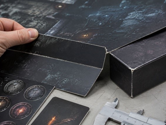 Close-up inspection of a dark mounted game board fold crack, white paper fiber, worn dark box corner, dark punchboard tokens, and caliper on a sample table